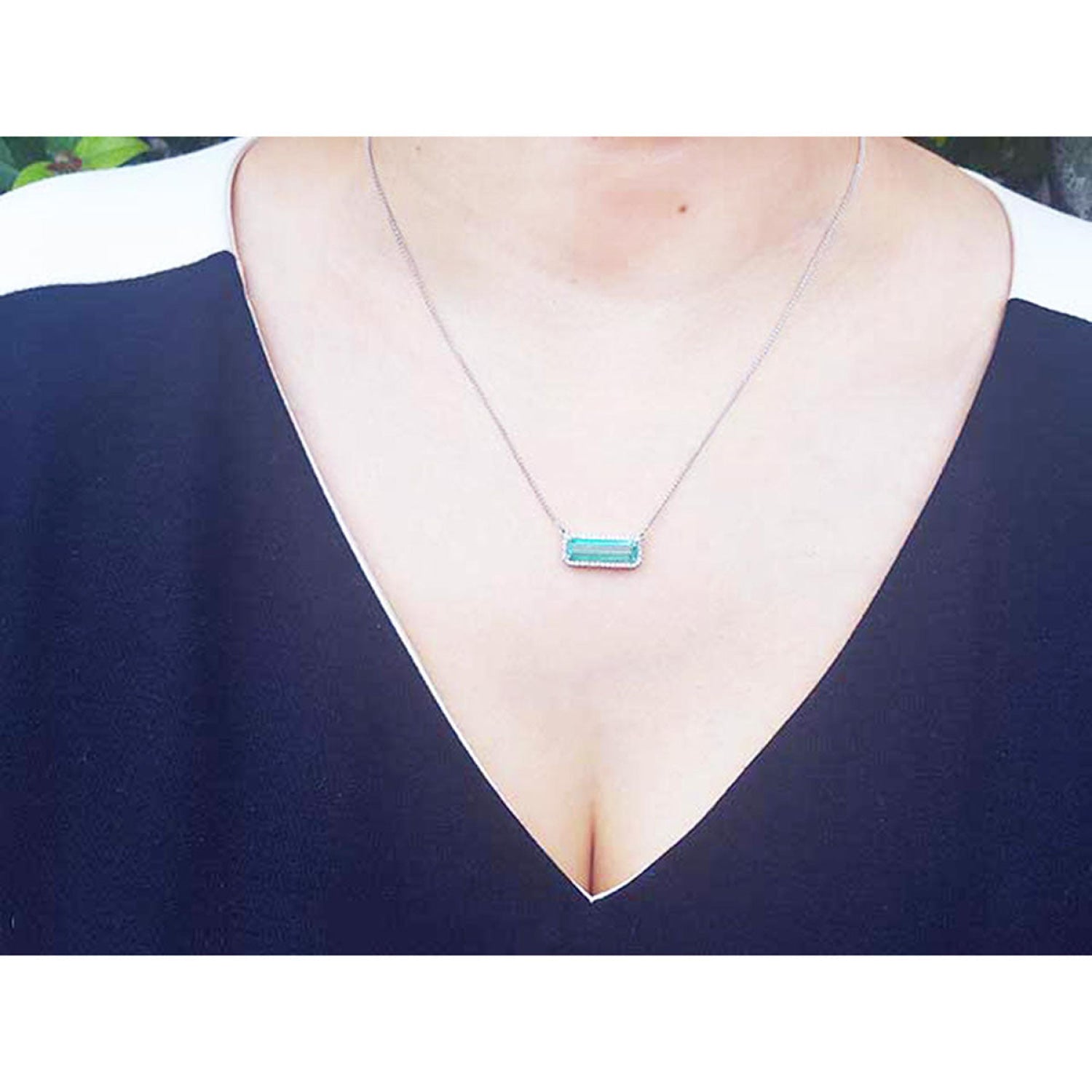 18K white gold necklace with a rectangular emerald-cut Colombian emerald and halo diamonds, worn by a model in a dark navy blue V-neck top.