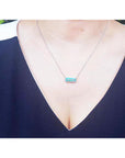 18K white gold necklace with a rectangular emerald-cut Colombian emerald and halo diamonds, worn by a model in a dark navy blue V-neck top.