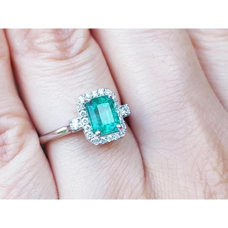 An inexpensive emerald engagement ring with a halo diamond white gold on a finger.