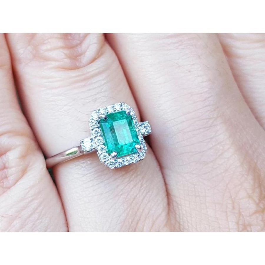 An inexpensive emerald engagement ring with a halo diamond white gold on a finger.