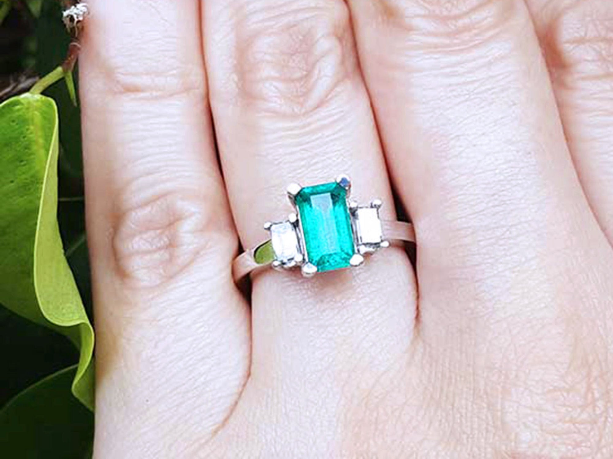 A Colombian emerald engagement ring on a finger against a natural background.
