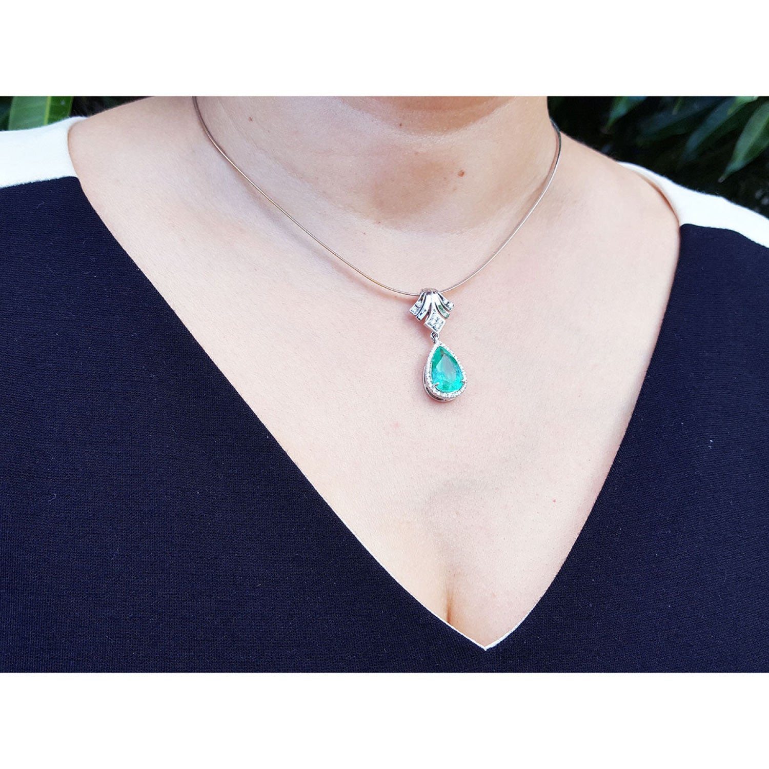 Pear-shaped emerald enhancer necklace with halo diamonds on an 18K gold chain, worn on a person.
