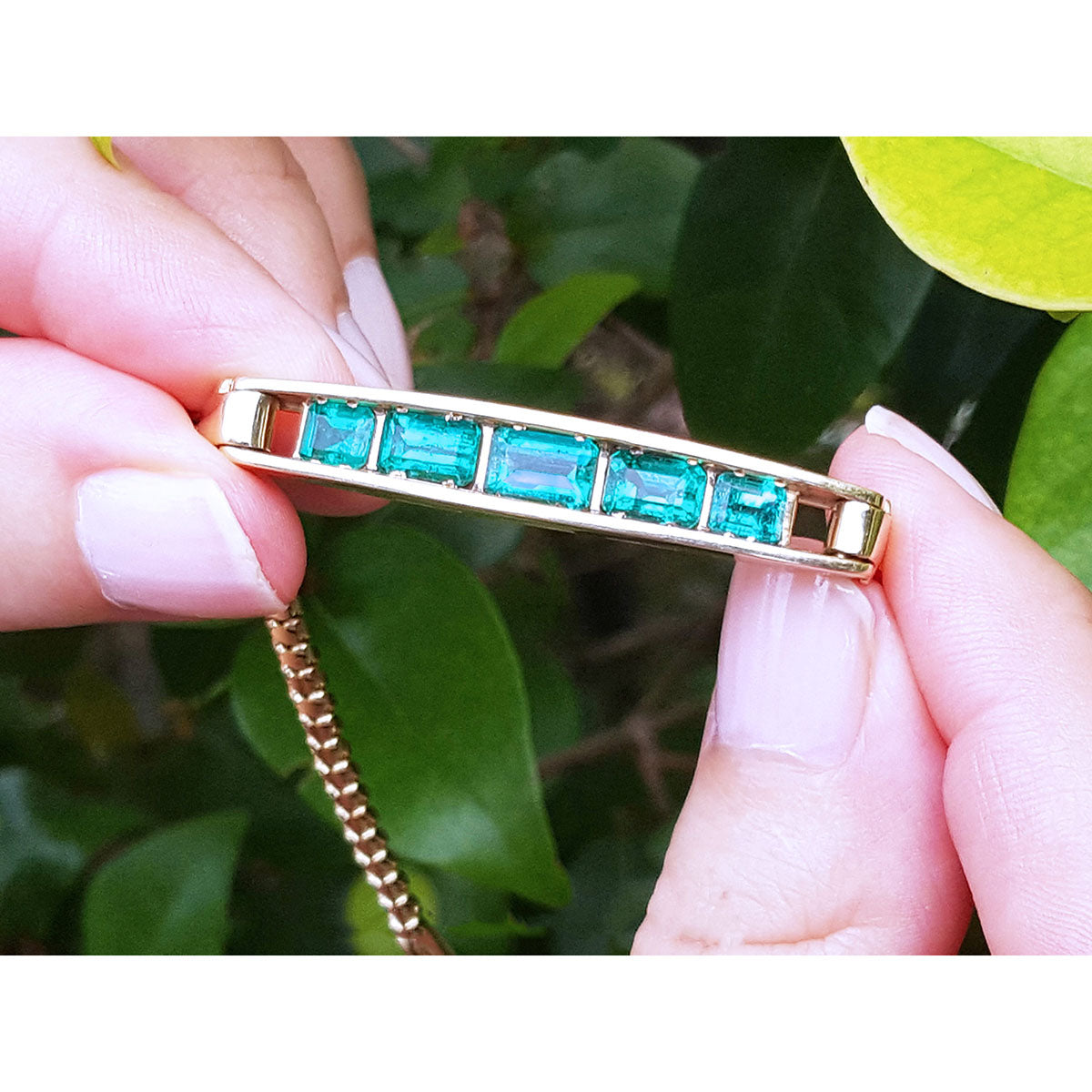 Gold bracelet with natural emeralds stones held by a hand against a green leafy background.