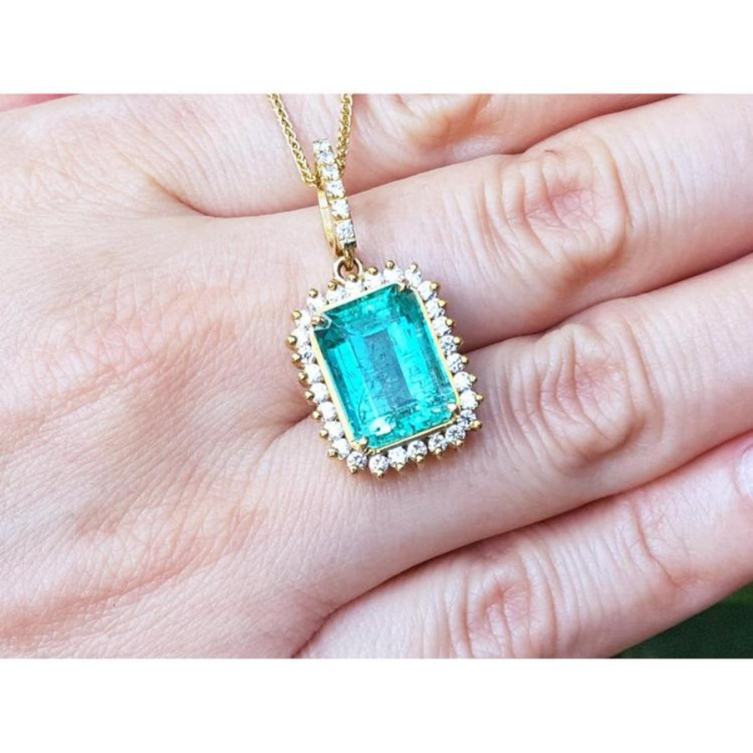 Emerald-cut emerald pendant with halo diamonds in 18K gold, worn on a hand.