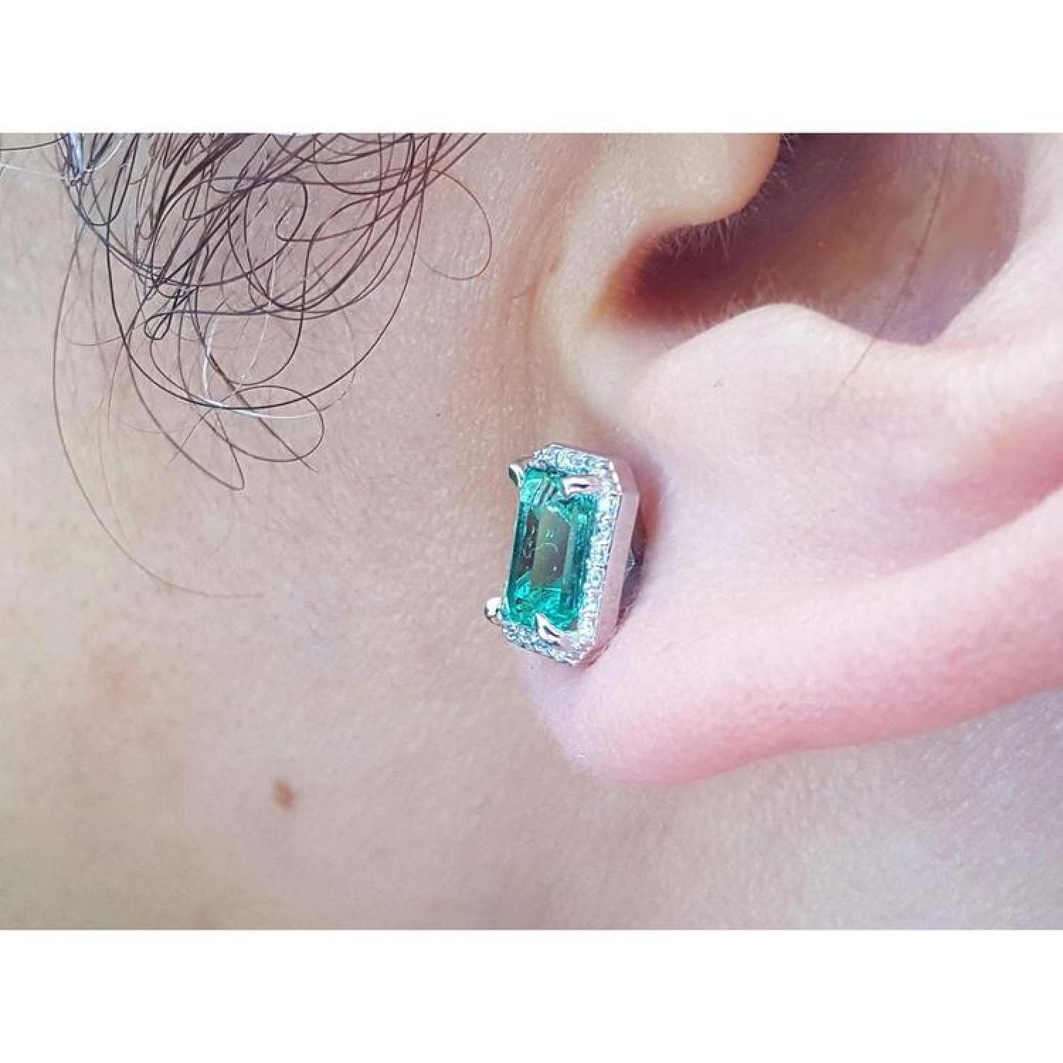 Emerald-cut natural emerald stud earring with halo diamonds in 14K white gold, worn on an ear.