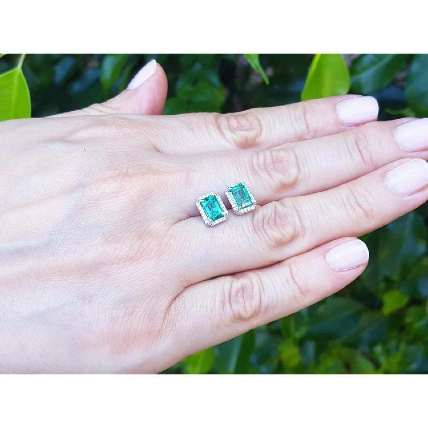 Emerald-cut natural Colombian emerald stud earrings with diamond halo, set in 14K white gold, on a hand.