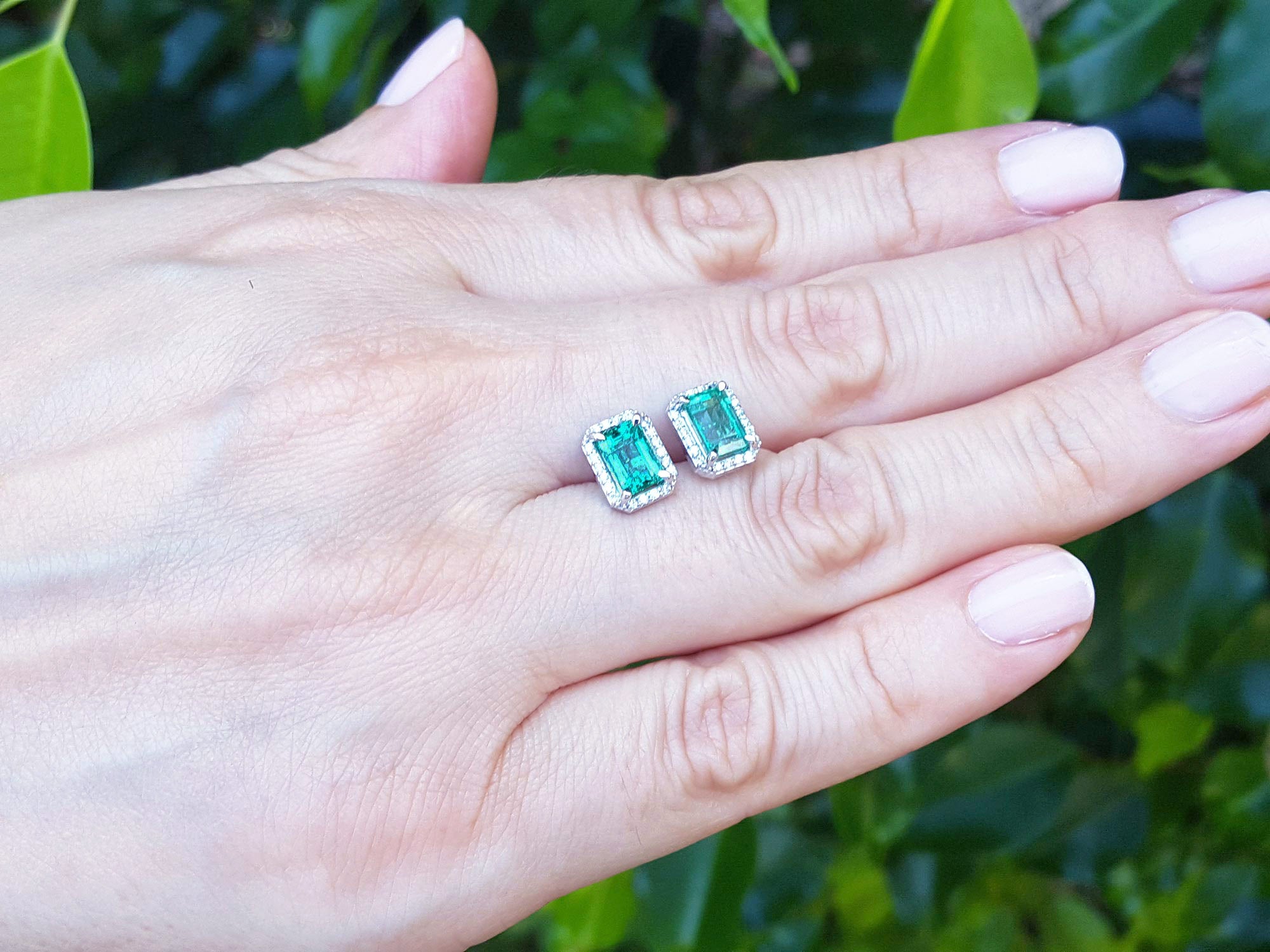 A pair of emerald and diamond Colombian emerald stud earrings on a hand with a blurred green background.