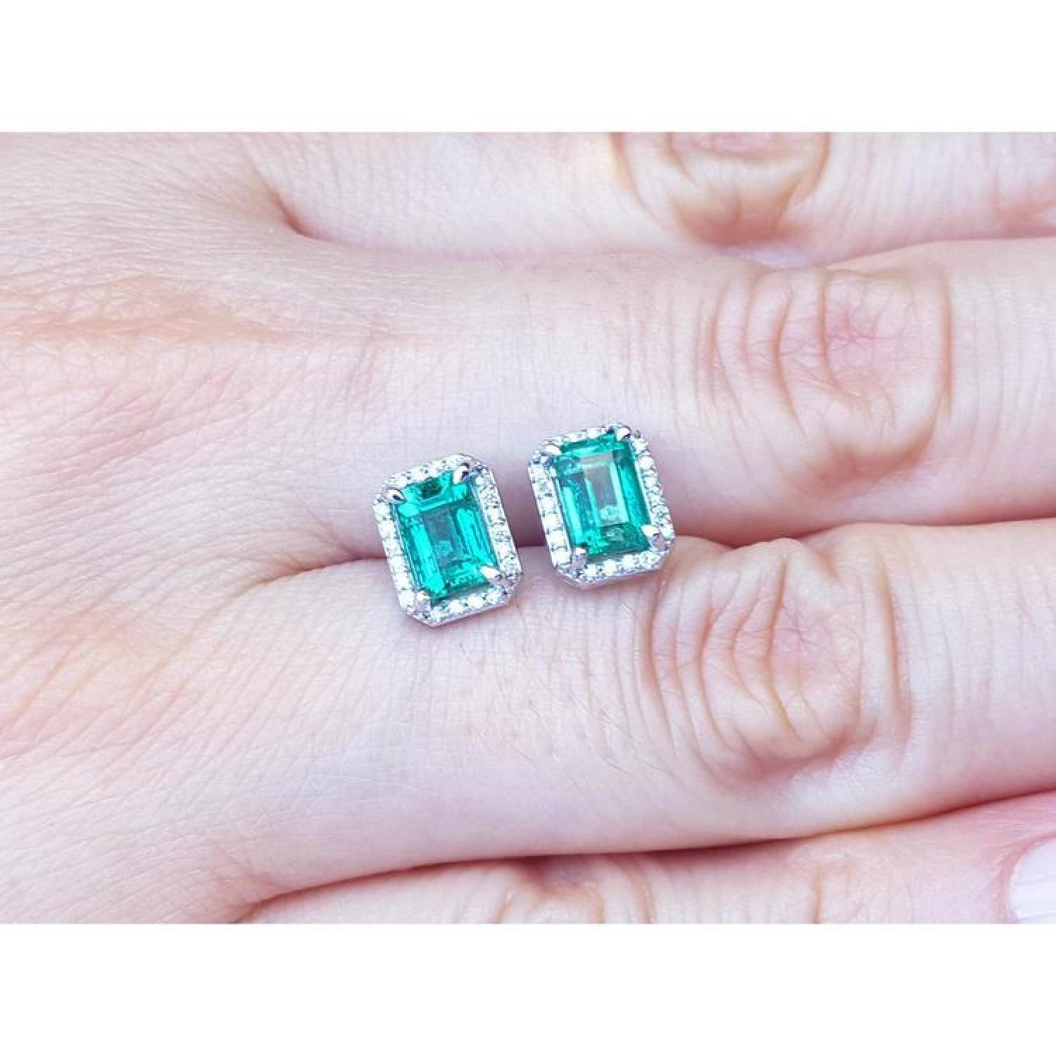 Emerald-cut natural Colombian emerald stud earrings with a halo of diamonds in 14K gold, held on a hand.