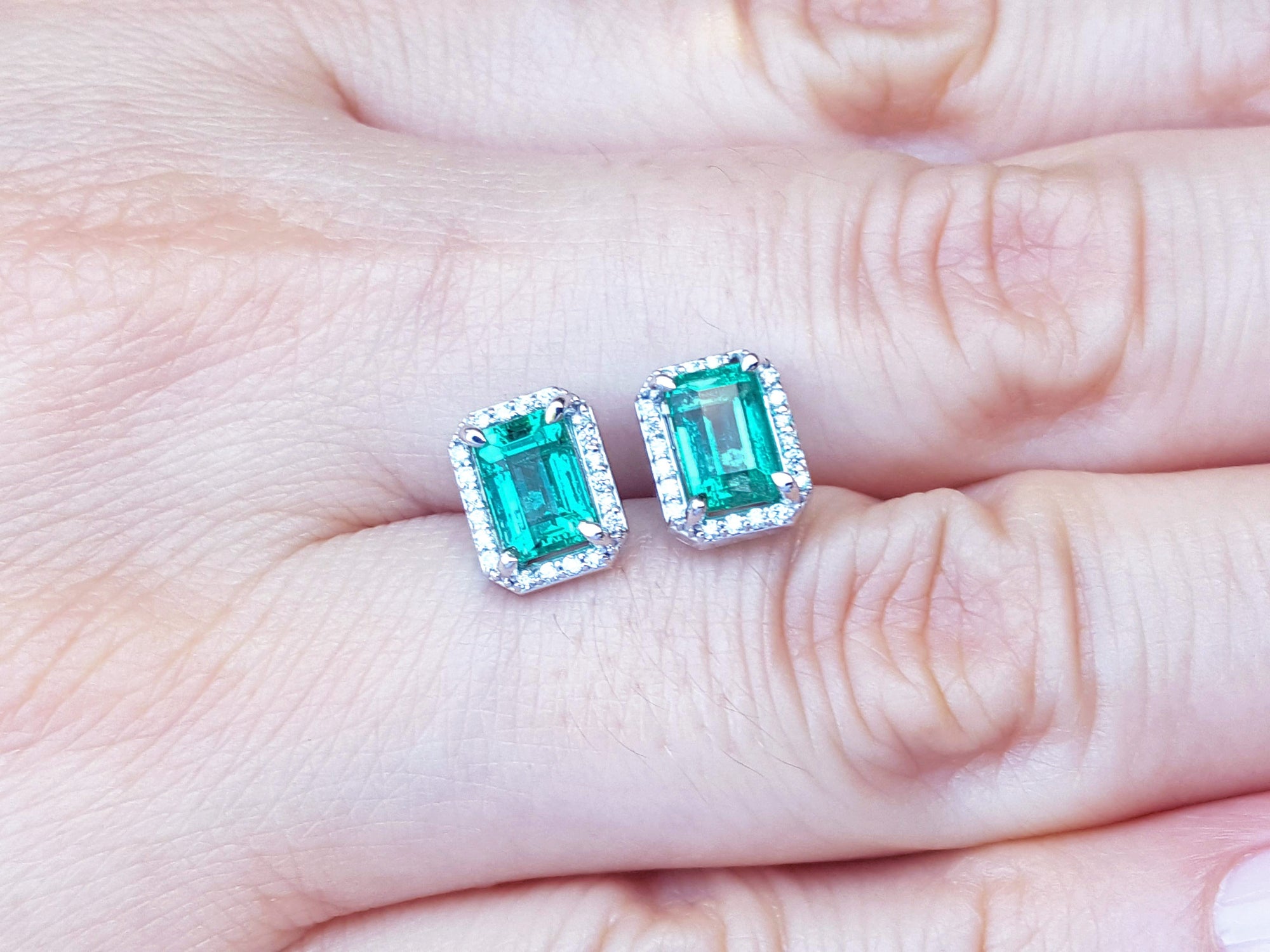 Emerald green emerald-cut gemstone earrings with diamond halo on a hand.