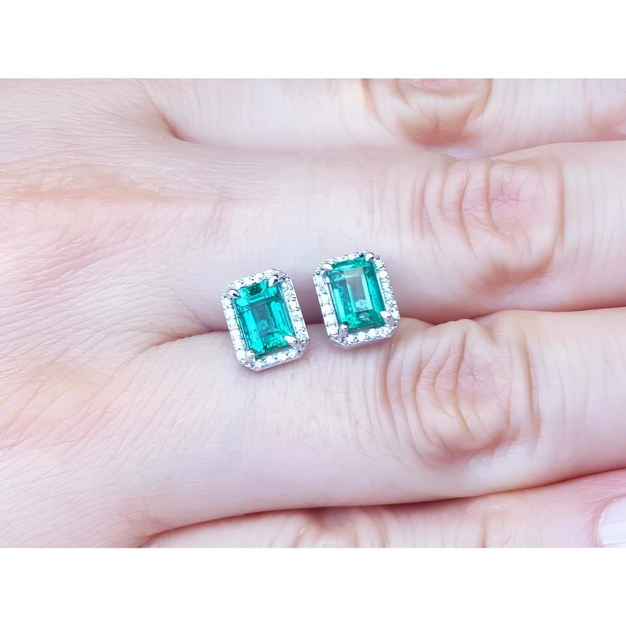 Emerald-cut natural Colombian emerald stud earrings with a halo of diamonds in 14K gold, held on a hand.