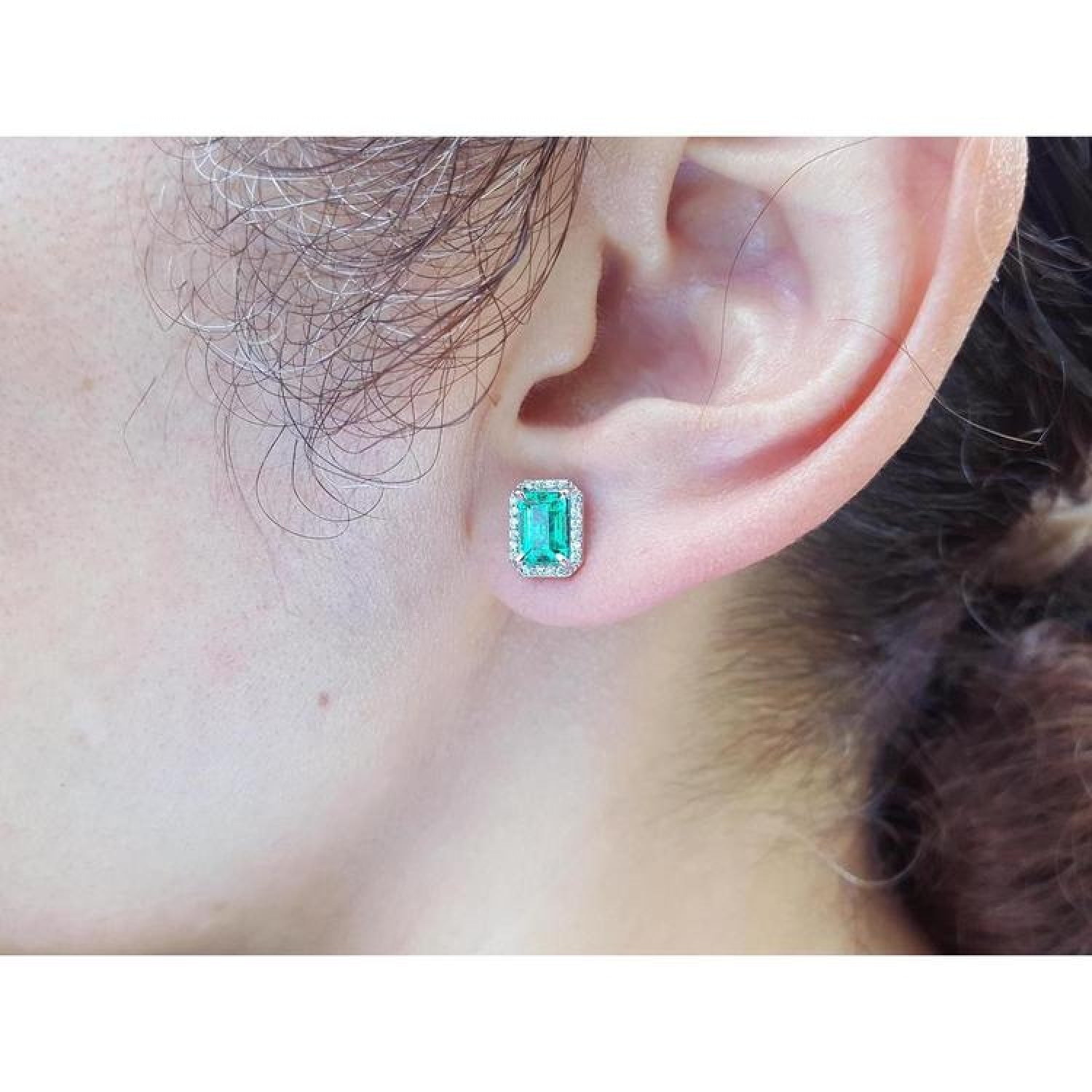 Emerald-cut natural emerald stud earrings with a halo of diamonds set in 14K gold on an ear.