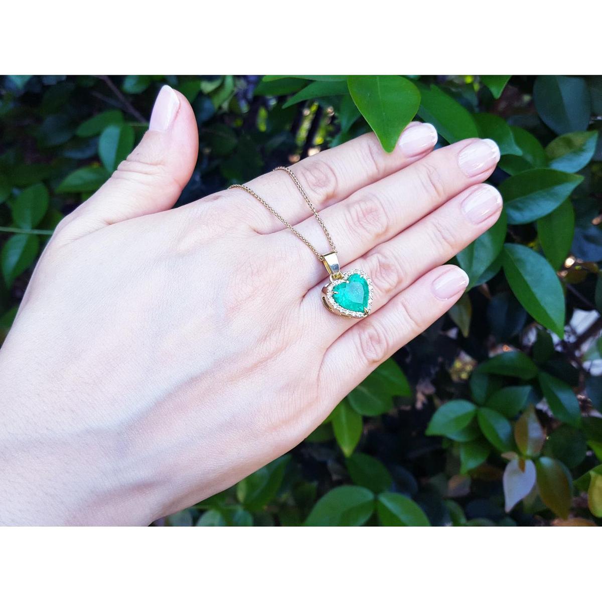 Hand holding a gold necklace with a natural emerald heart pendant against a leafy background.