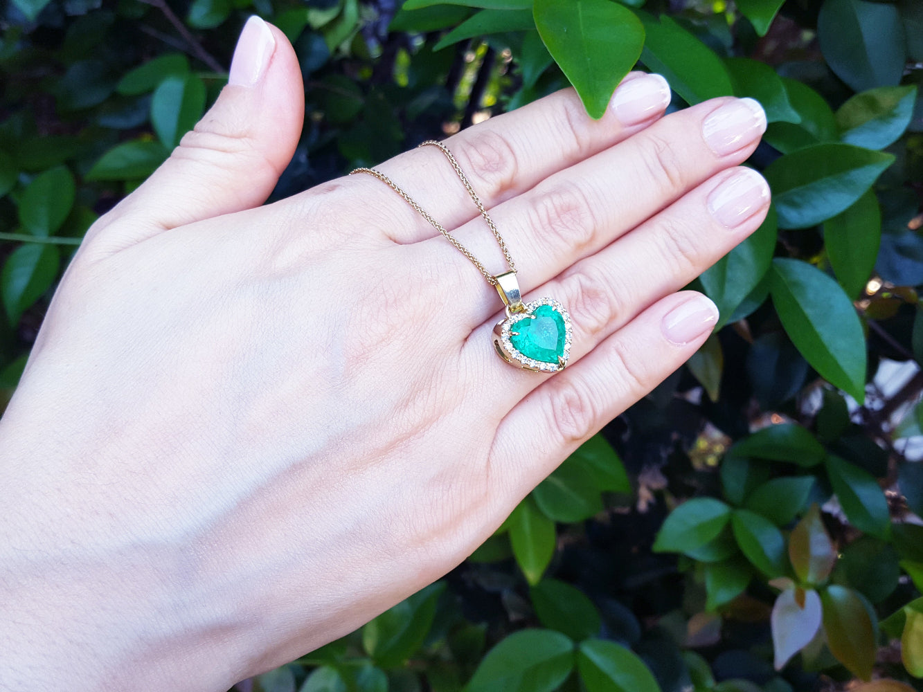 Hand holding a gold necklace with a natural emerald heart pendant against a leafy background.