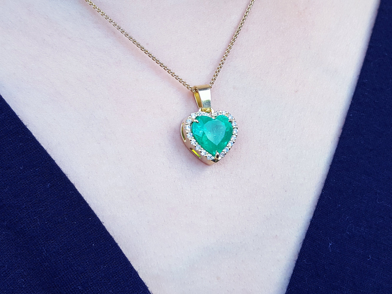 Heart-shaped emerald pendant on a gold chain against a white and blue fabric background.