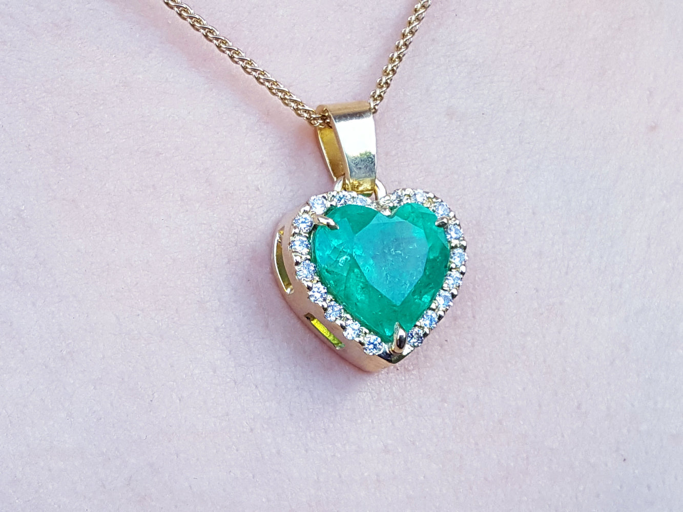 Heart-shaped emerald pendant with gold frame and diamonds on a light pink skin background.