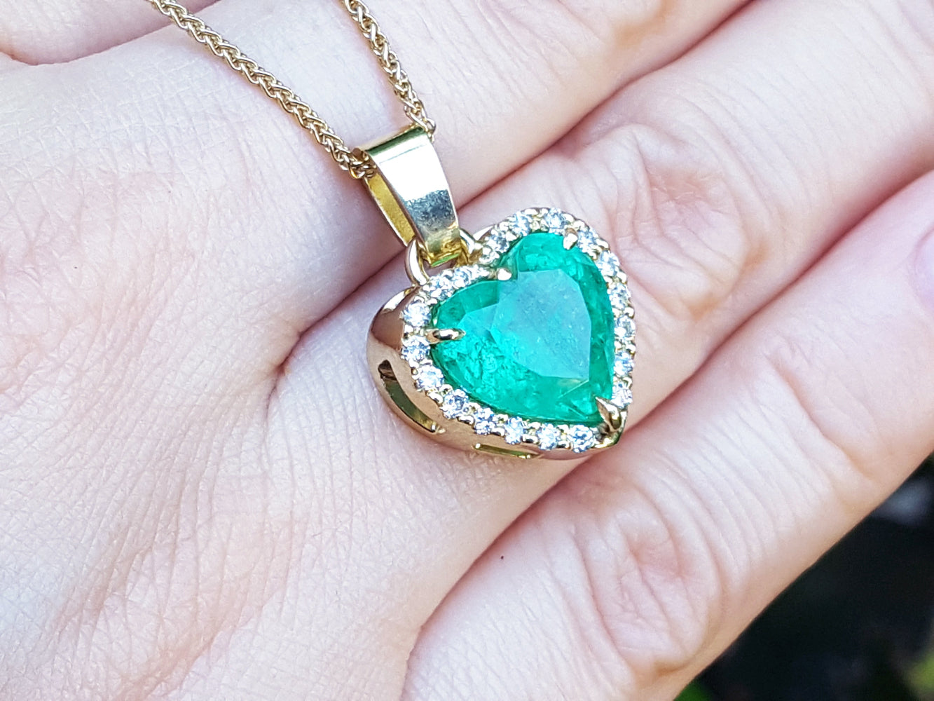 Heart-shaped emerald pendant on a gold chain held on a hand.