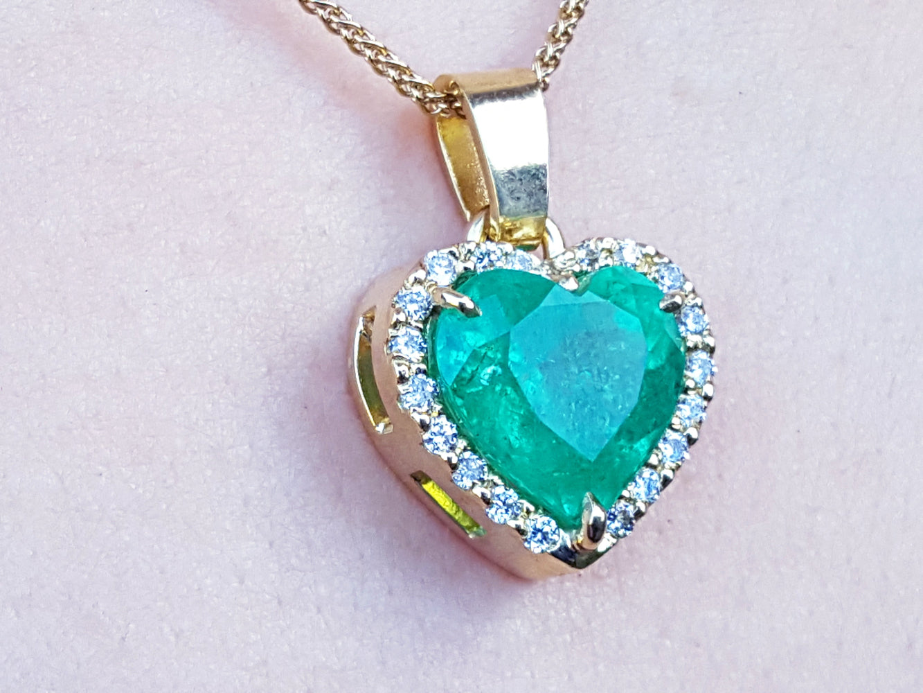 Heart-shaped emerald pendant with gold frame and diamonds on a light pink background.