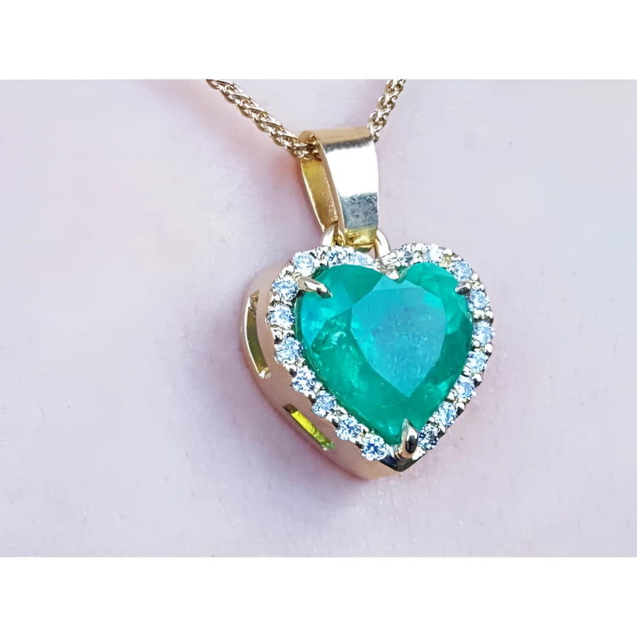 Heart-shaped emerald pendant with gold frame and diamonds on a light pink background.