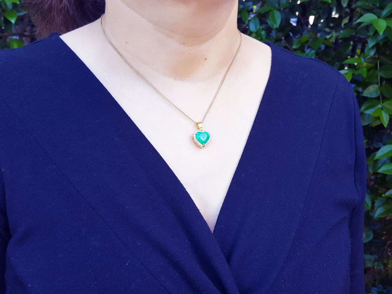A woman wearing a blue top with a necklace featuring a natural colombian emerald heart pendant.