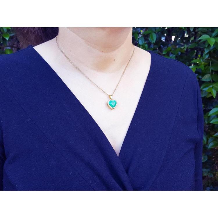 Heart-shaped emerald pendant with diamond halo on 18K yellow gold chain worn on a woman.