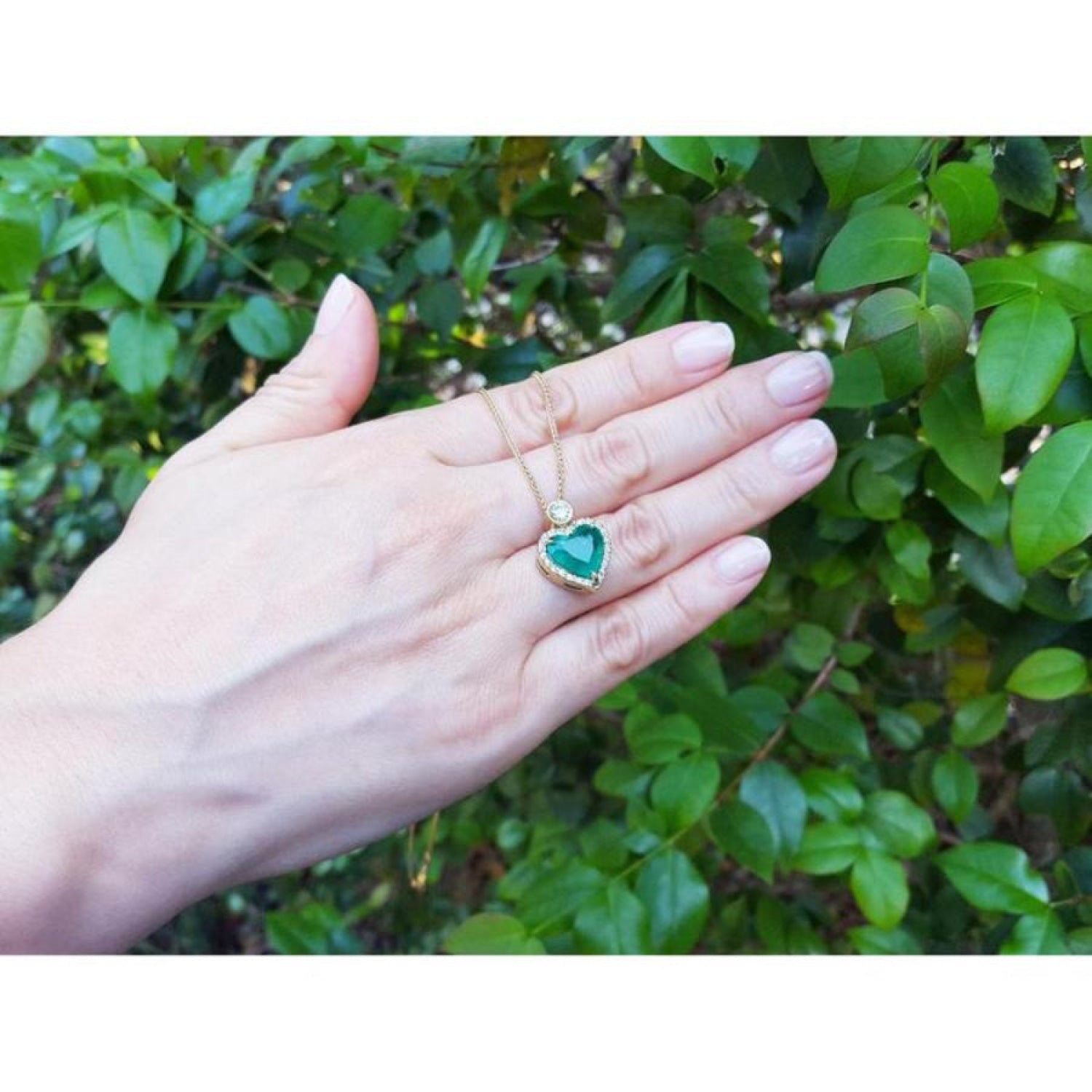 Emerald heart pendant with diamond halo on an 18k gold chain, held against green foliage.