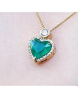 Heart-shaped Muzo colombian emerald pendant with gold chain on a light pink background.