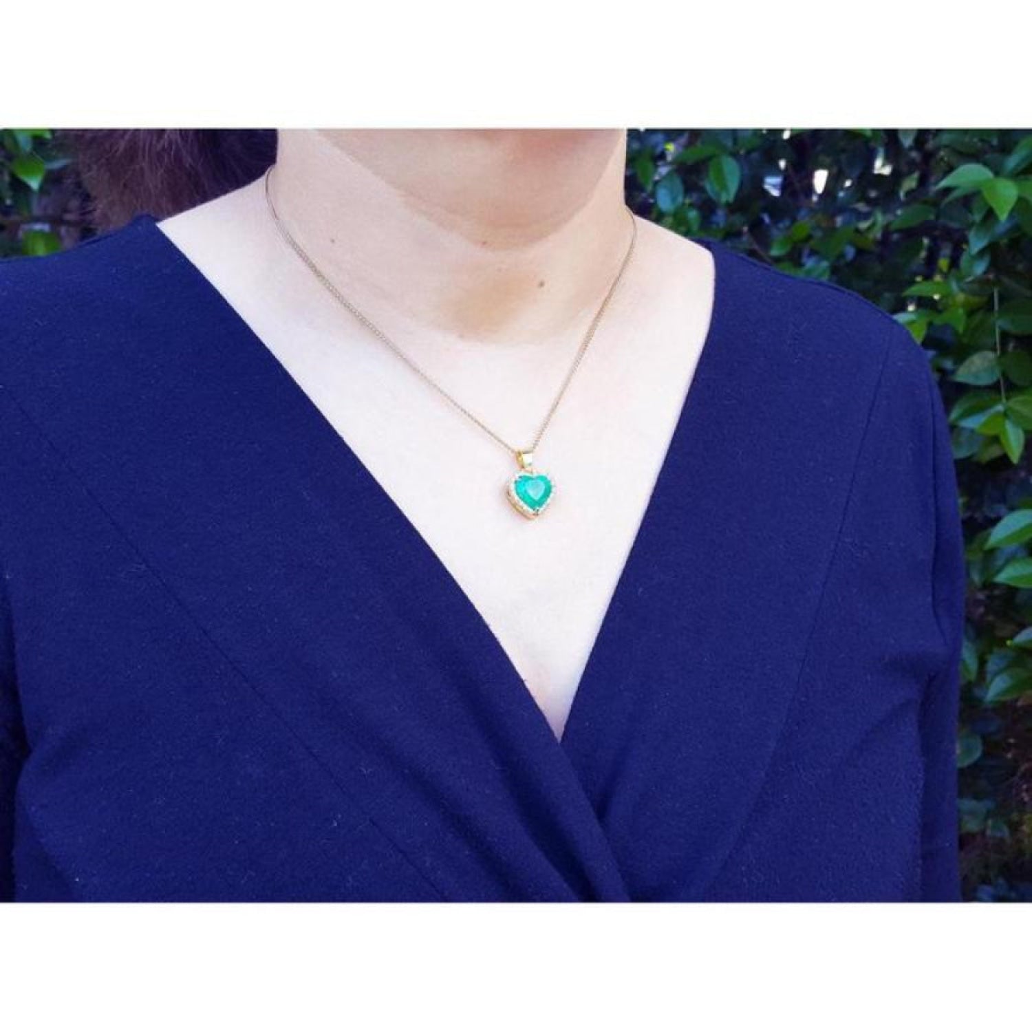 Heart-shaped emerald pendant with diamond halo on 18K yellow gold chain worn on a woman.