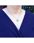 Heart-shaped emerald pendant with diamond halo on 18K yellow gold chain worn on a woman.