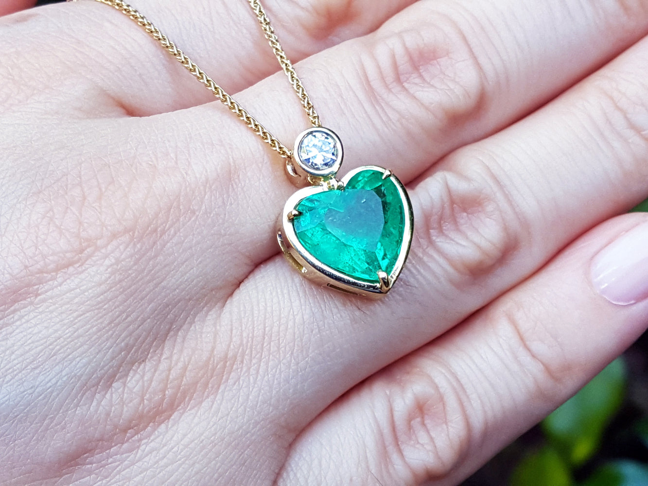 Heart-shaped emerald pendant on a gold chain held on a hand.