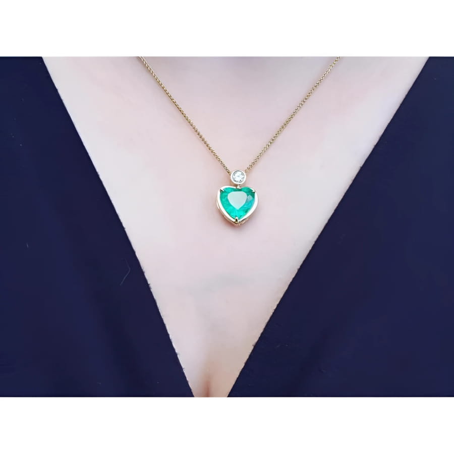 Heart-shaped emerald pendant necklace in 18K yellow gold with a diamond accent on a woman's neck.