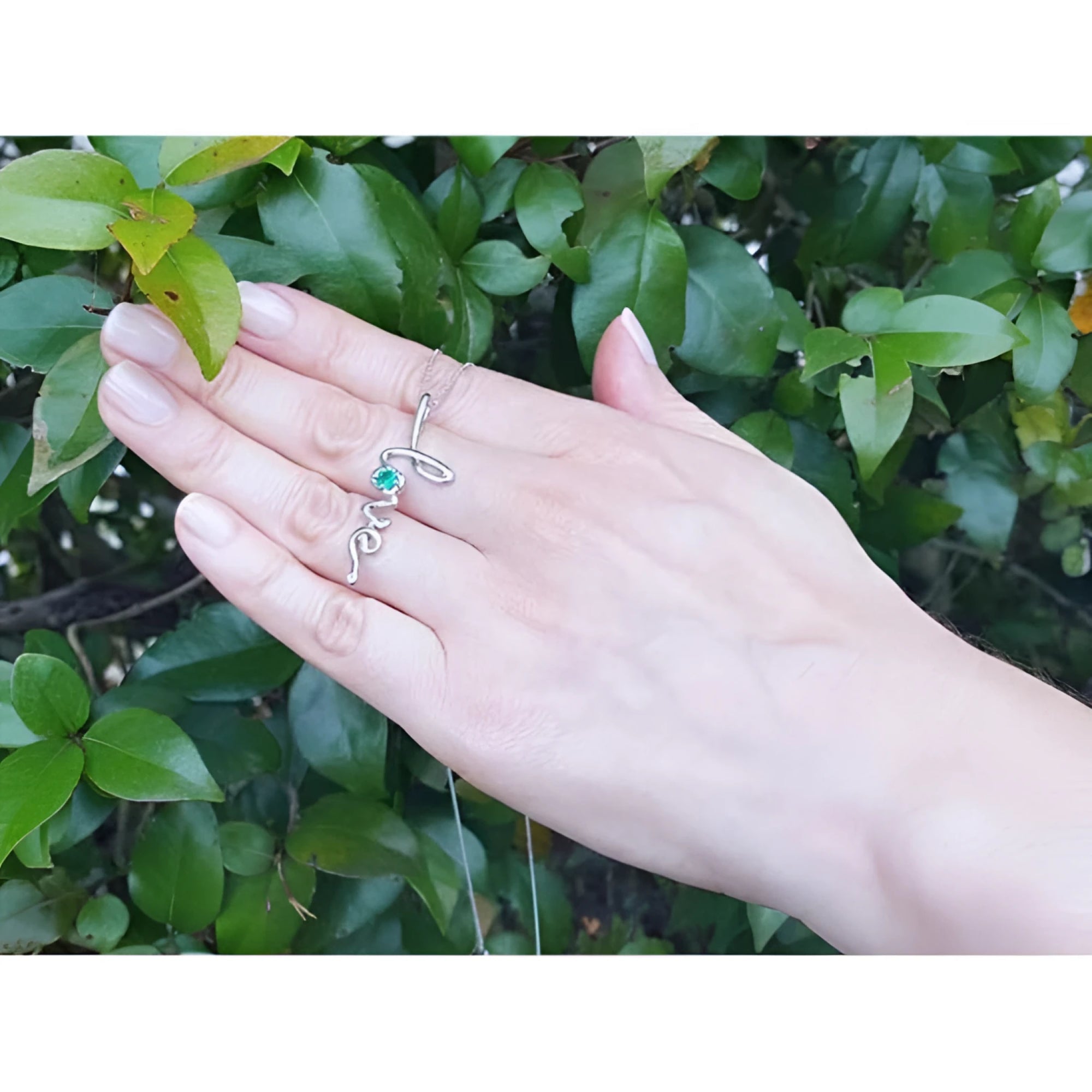 14K white gold "love" script pendant necklace with a round Colombian emerald, worn on a hand.