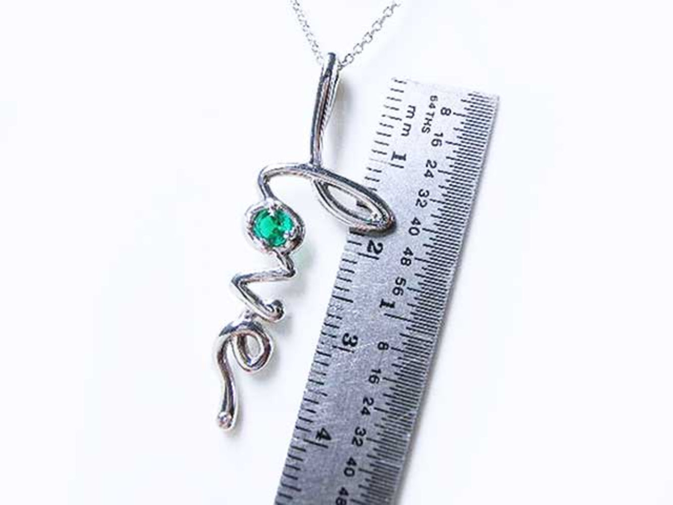 A white gold necklace with a round cut emerald love pendant next to a ruler for size reference.