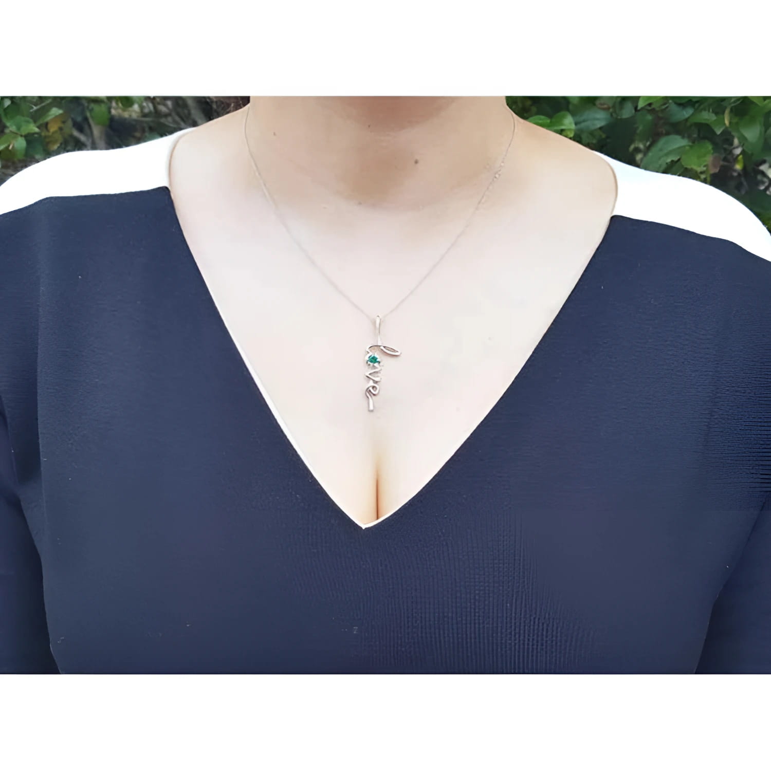 14K white gold "Love" script pendant with a round Colombian emerald, worn on a woman's neck.