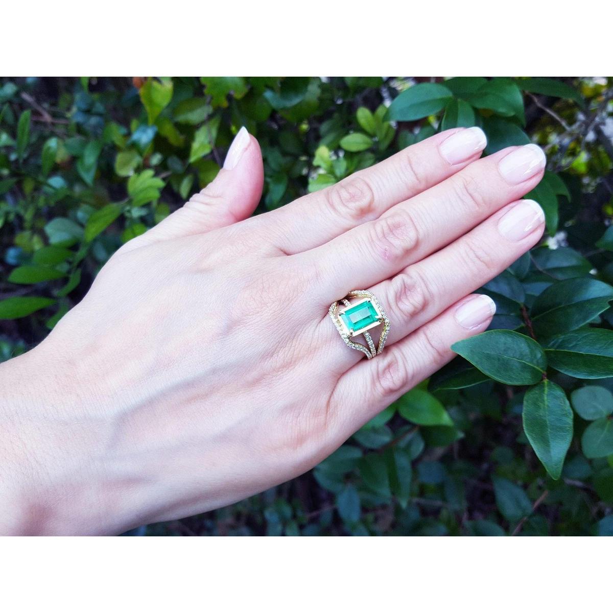 Hand wearing a 18k yellow gold emerald ring for women against a leafy background.