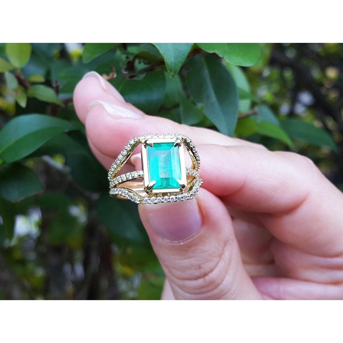 A Muzo mine Colombian emerald ring for women held by a hand against a blurred green leafy background.