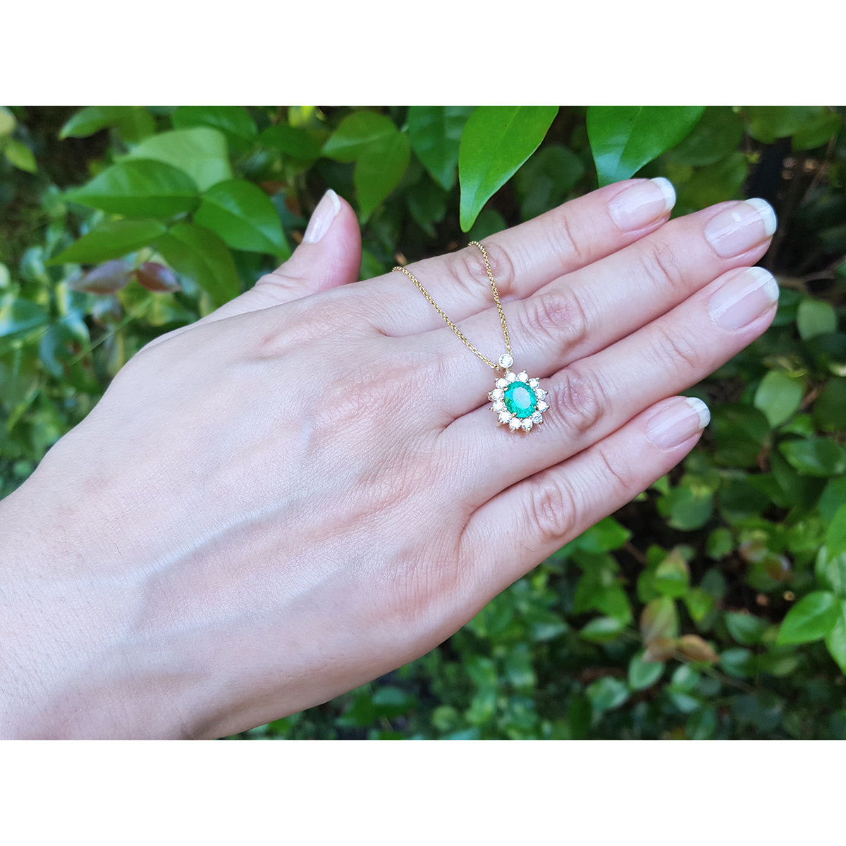 Hand displaying a yellow gold necklace with a oval cut emerald pendant against a green leafy background