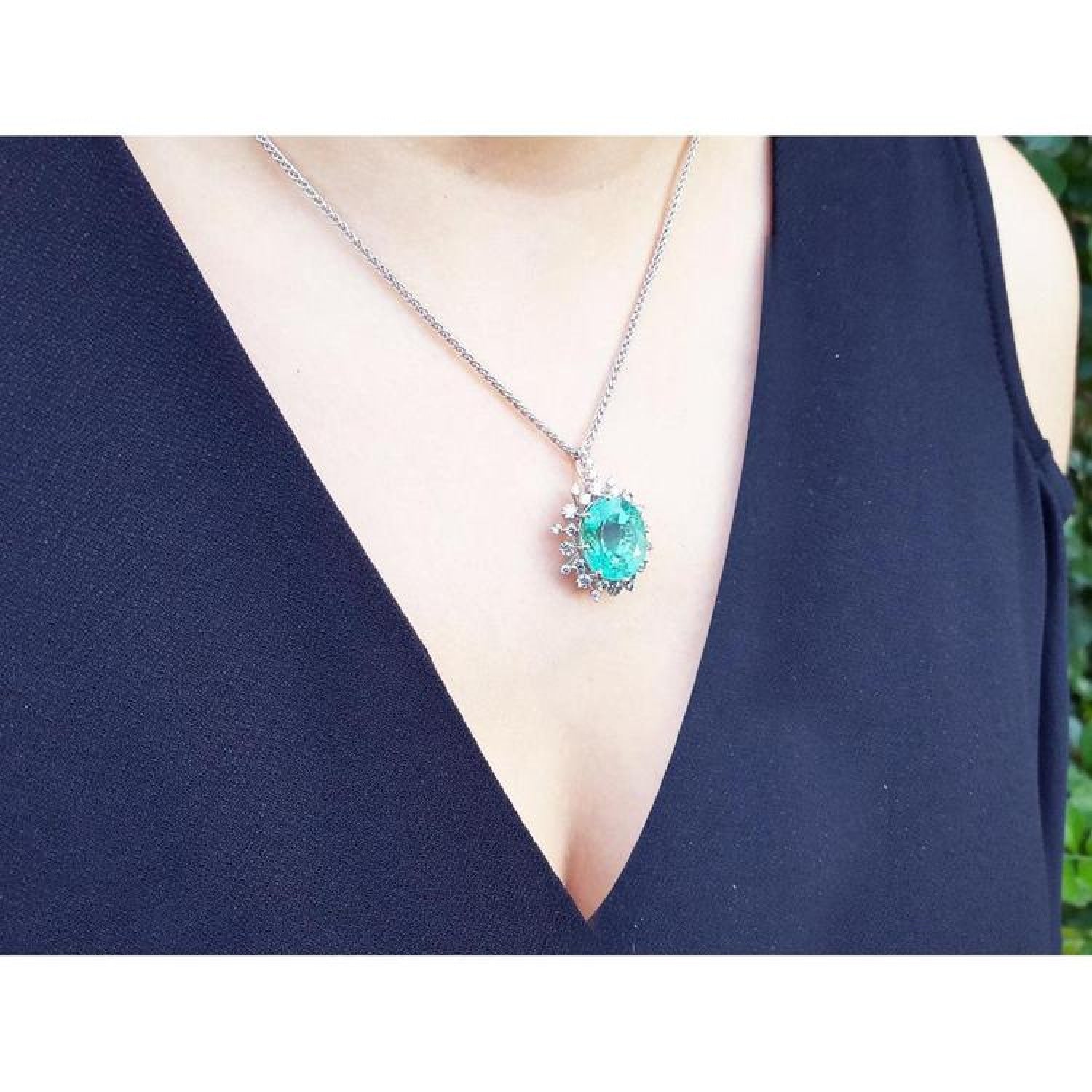 Oval cut emerald pendant with halo diamonds on a 14K white gold chain, worn on a woman's chest.