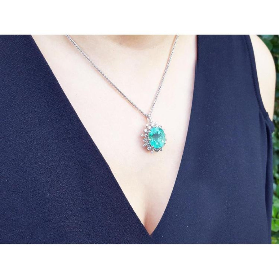 Oval cut emerald pendant with halo diamonds on a 14K white gold chain, worn on a woman's chest.