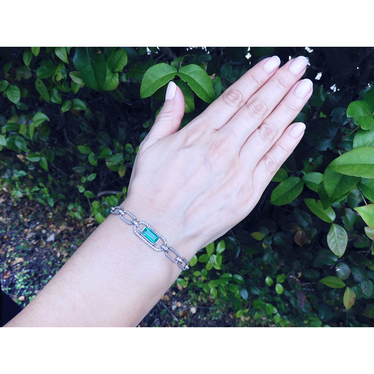 A lady&#39;s hand wearing a bracelet with a colombian emerald stone against a green leafy background.