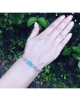 A lady's hand wearing a bracelet with a colombian emerald stone against a green leafy background.