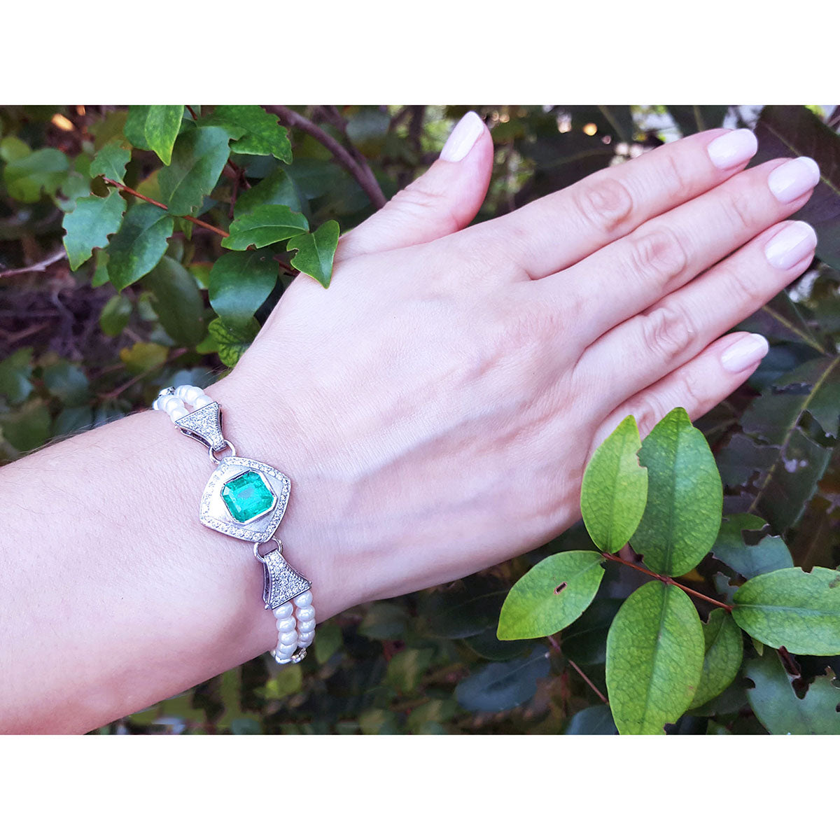 Hand wearing a pearl bracelet with a square emerald against a leafy background.