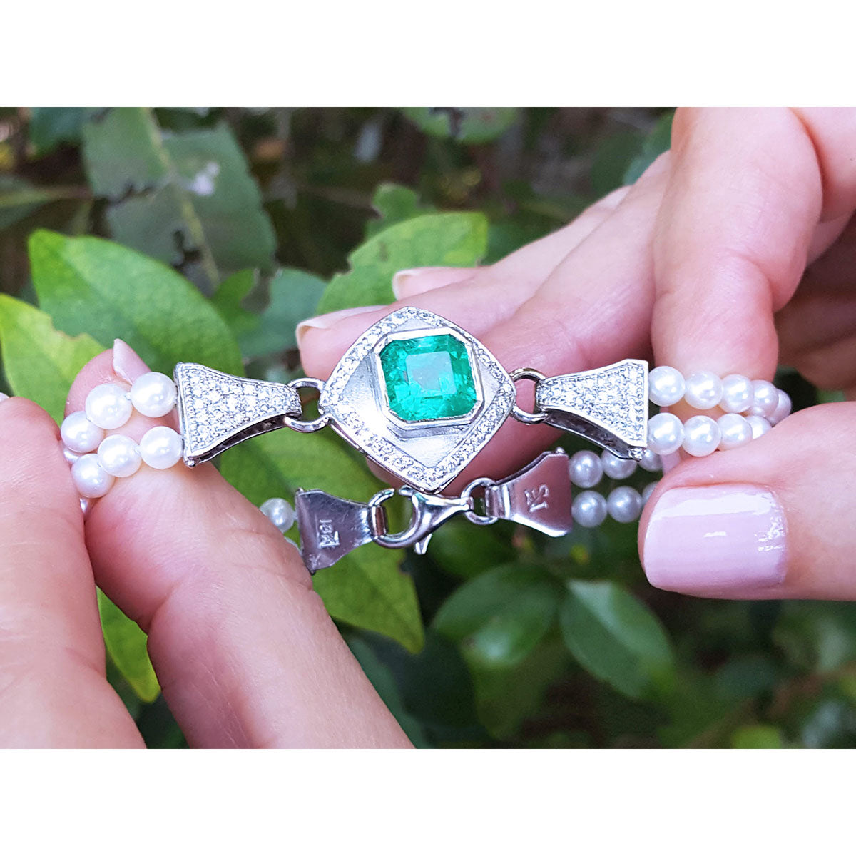 Pearl bracelet with a square colombian emerald held by hands against a leafy background.