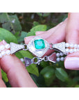Pearl bracelet with a square colombian emerald held by hands against a leafy background.