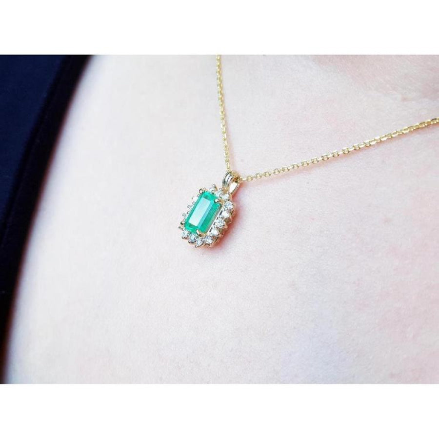 14K yellow gold pendant with a rectangular emerald surrounded by a halo of natural diamonds, worn on a neck.