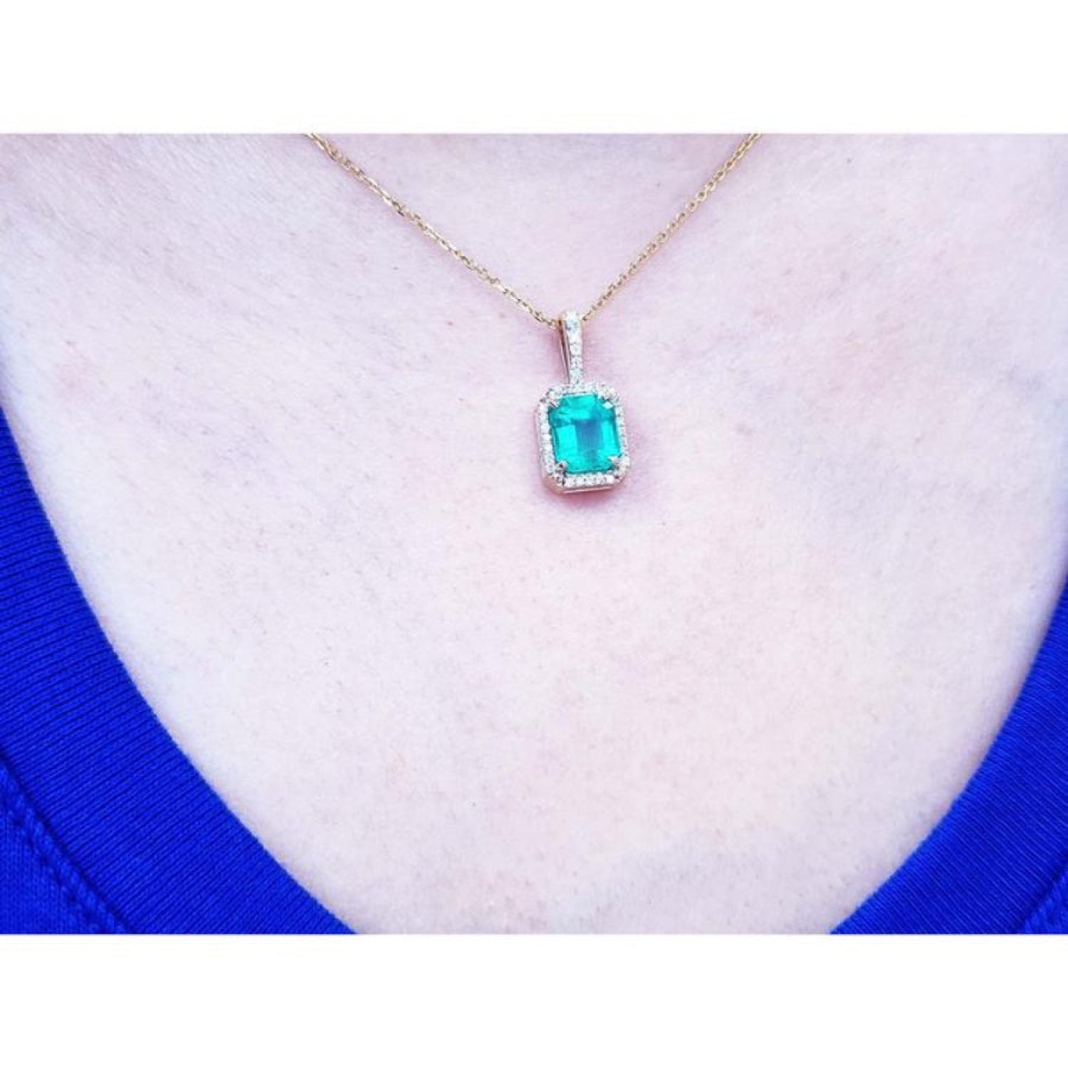 Emerald-cut Colombian emerald pendant with diamond halo on a 14k yellow gold chain, worn on a neck.