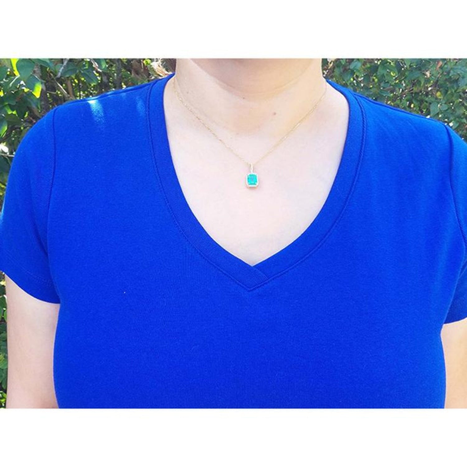 Emerald-cut emerald pendant with diamond halo on a thin gold chain, worn on a blue shirt.