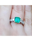 Hand wearing a square emerald ring with side diamond princess cut in white gold.