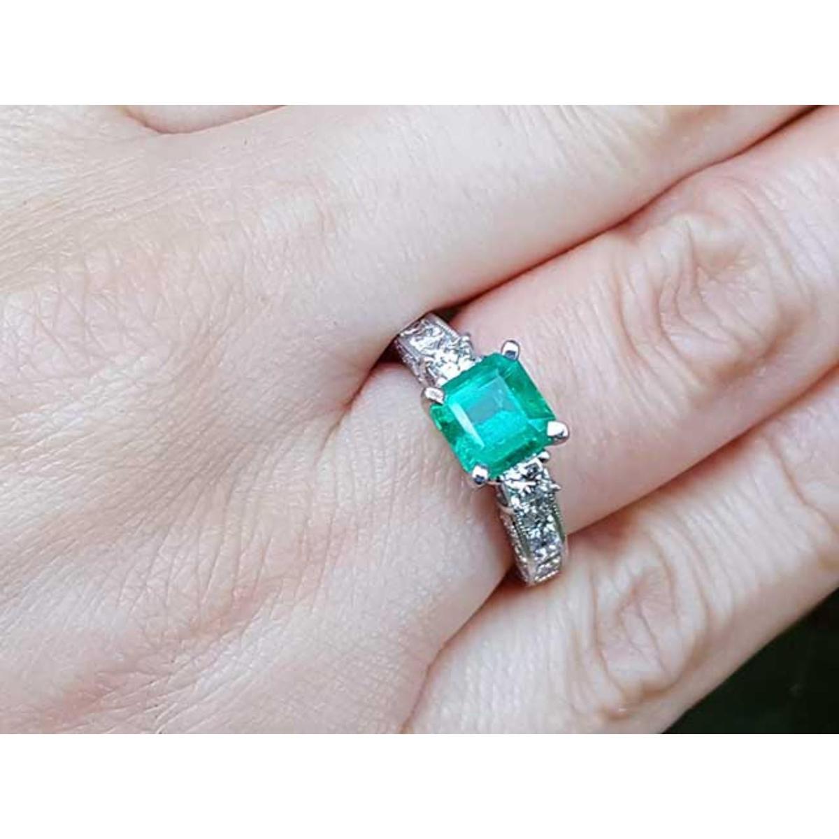 A square emerald engagement ring with princess cut side diamonds on a person's hand.