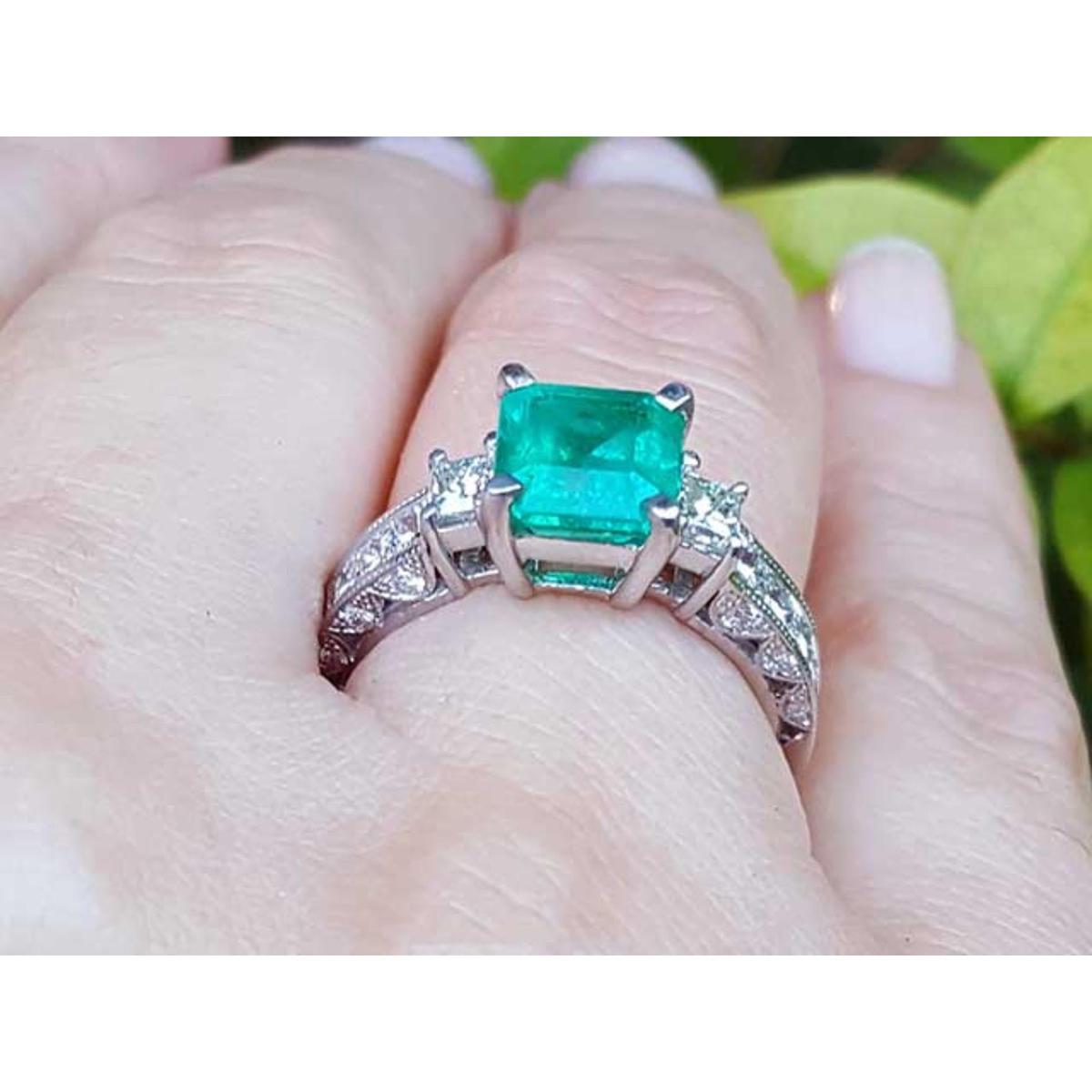 On a woman's hand a square Colombian emerald engagement ring, against a leafy background.