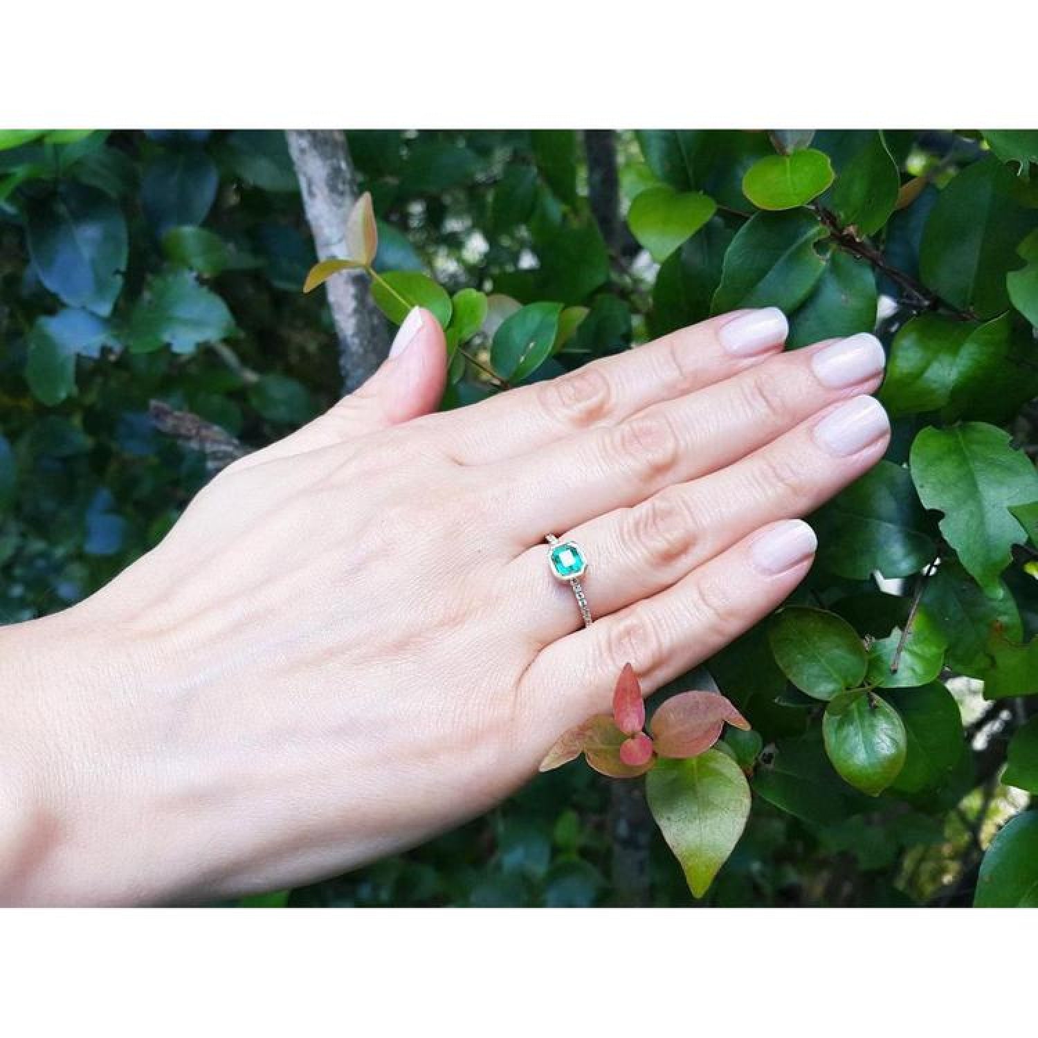 Bezel-set emerald ring with accent diamonds in 14K yellow gold, worn on a hand amidst green leaves.